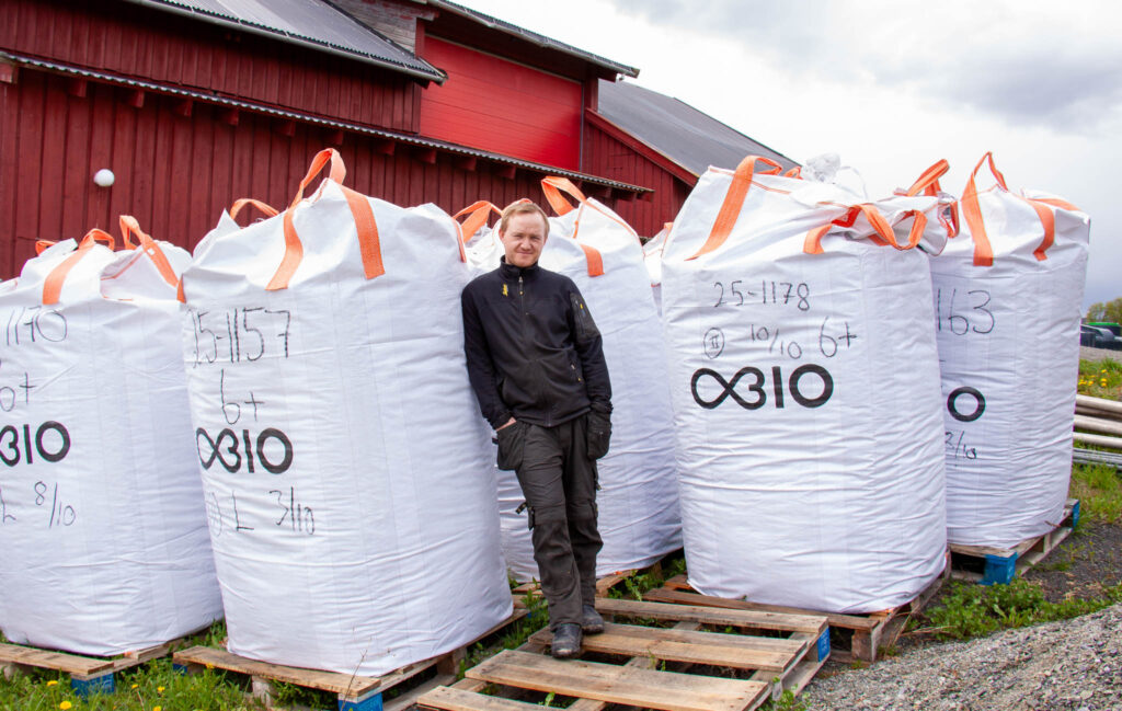 _MG_2688 – Obio AS Man in a black jacket stands among large white bulk bags with orange straps outside a red warehouse building.
